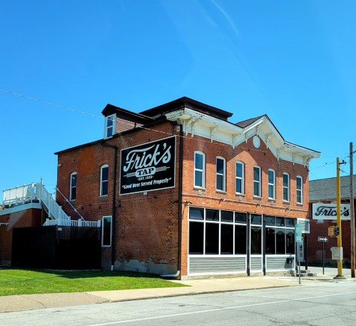 A brick building with a sign reading "Frick's Tap" and a slogan, under a clear blue sky.