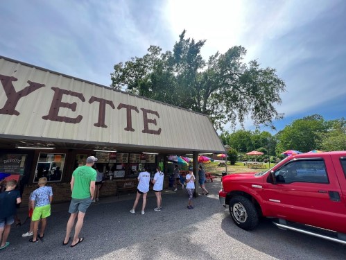 A group of people gathers outside a food stand named "Yette," with a red truck parked nearby and trees in the background.