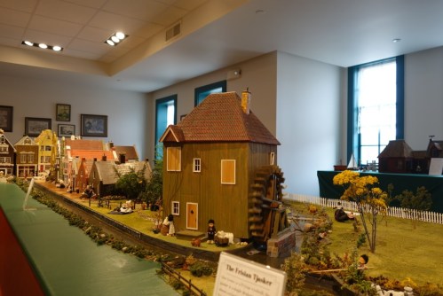 A detailed model village with a wooden house, windmill, and miniature figures, displayed in a bright room.