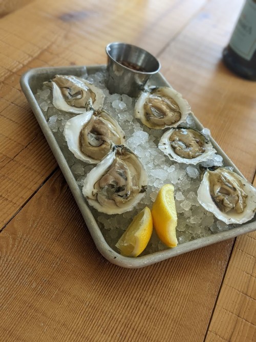 A metal tray filled with ice, featuring six oysters on the half shell and two lemon wedges.