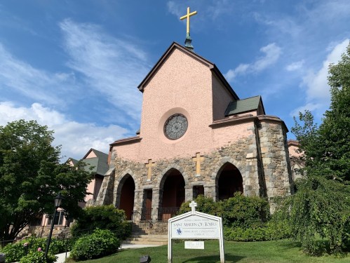 A pink church with a stone base, large round window, and a cross on top, surrounded by greenery and a sign in front.