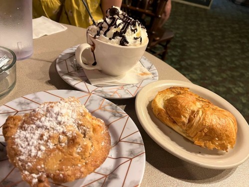 A plate with a powdered muffin, a cup of ice cream topped with chocolate sauce, and a croissant on a table.