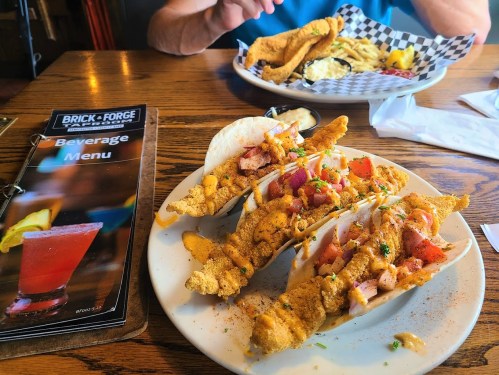 A plate of fish tacos topped with diced tomatoes and sauce, alongside a beverage menu and a plate of fries in the background.