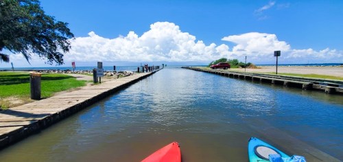 A serene waterway leads to a beach, with kayaks in the foreground and fluffy clouds in a blue sky above.