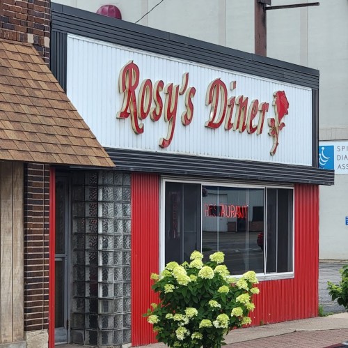 Sign for "Rosy's Diner" with a red and white exterior, featuring a glass block wall and a flower bush in front.