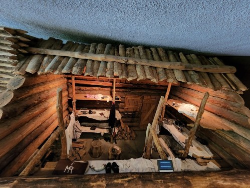 A rustic wooden cabin interior with bunks, clothing, and various items arranged on shelves and the floor.
