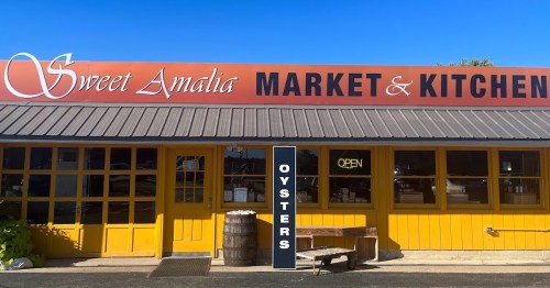 Exterior of Sweet Amalia Market & Kitchen, featuring a bright yellow facade and a sign for oysters. Clear blue sky above.