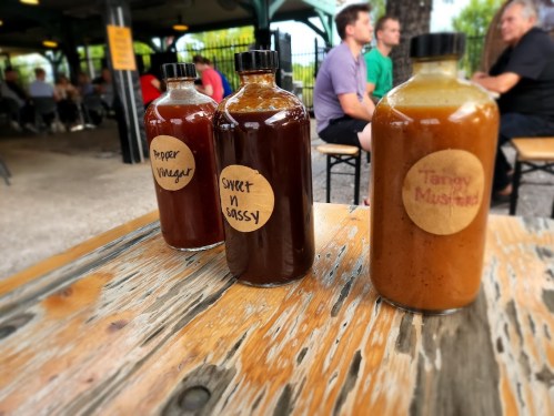 Three bottles of sauce labeled "Hoper Vinger," "Sweet 'n Sassy," and "Tangy Mustard" on a wooden table.