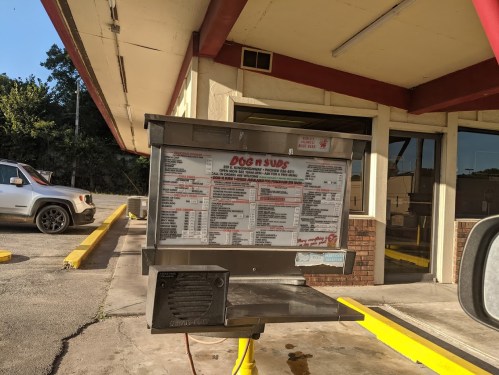 Menu board for a drive-thru restaurant featuring hot dogs and subs, with prices and options listed.