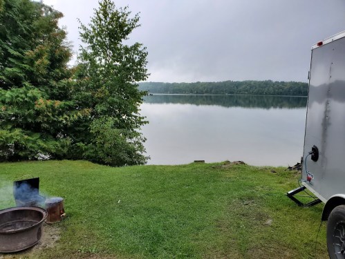 A serene lakeside view with a grassy area, a fire pit, and a trailer beside calm waters under a cloudy sky.