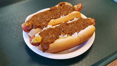 Two hot dogs in buns topped with chili, mustard, and chopped onions on a white plate.