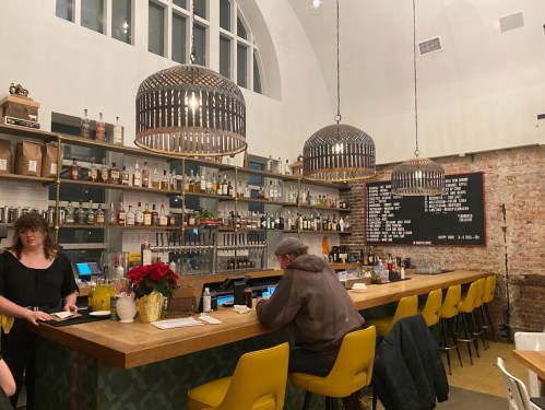 A cozy bar with a well-stocked shelf, warm lighting, and patrons seated at a wooden counter.