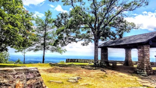 Scenic view from a stone pavilion surrounded by trees, overlooking a distant landscape under a blue sky.