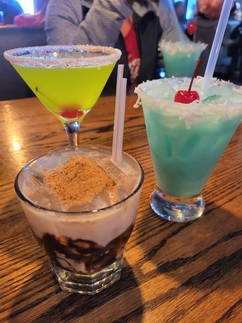 Three colorful cocktails on a wooden table: a green drink, a blue drink with a cherry, and a layered chocolate drink.