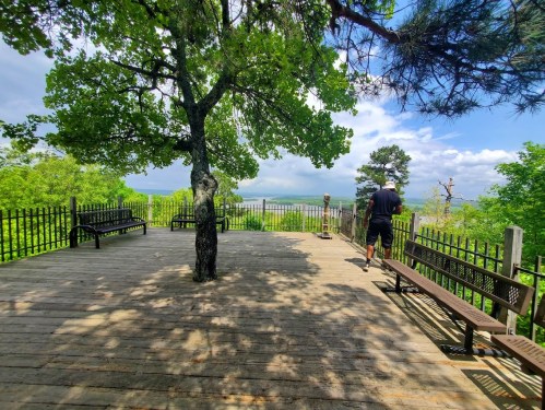 A wooden observation deck surrounded by trees, with a person walking away and a scenic view in the background.