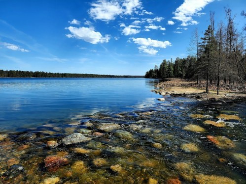A serene lake scene with clear blue water, colorful rocks, and a backdrop of trees under a bright sky with fluffy clouds.