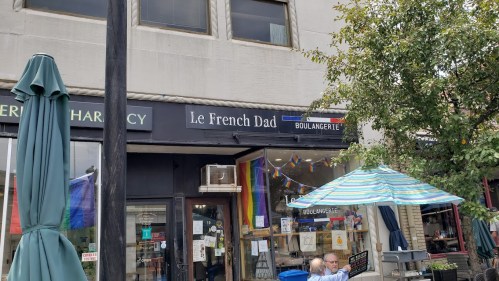 A storefront labeled "Le French Dad Boulangerie" with rainbow flags and outdoor seating under an umbrella.
