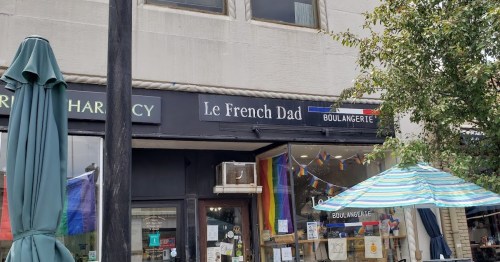 A storefront with a sign reading "Le French Dad Boulangerie," featuring rainbow decorations and an outdoor umbrella.