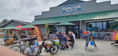 A bustling brewery with outdoor seating, umbrellas, and people enjoying drinks, bikes parked nearby.