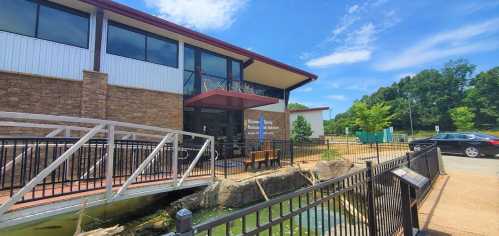 A modern building with large windows, a ramp, and a pond in front, surrounded by greenery and a clear blue sky.