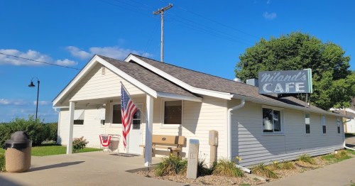A small, white café building with an American flag, a sign reading "Milan's Café," and a clear blue sky.