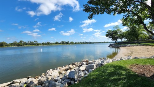 A serene lake view with clear blue skies, green trees, and a rocky shoreline under soft, fluffy clouds.
