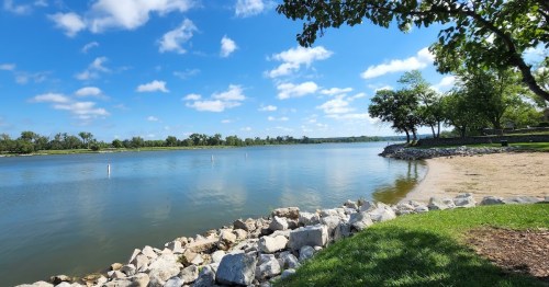 A serene lakeside view with clear blue skies, green trees, and a rocky shoreline.