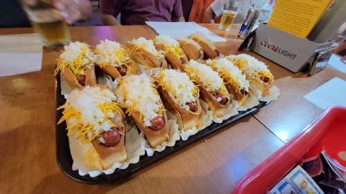 A tray of hot dogs topped with shredded cheese and diced onions, served in a casual dining setting.