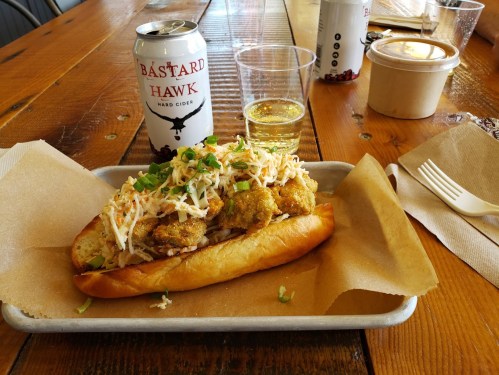 A loaded hot dog topped with fried pickles and slaw, served with a can of cider on a wooden table.