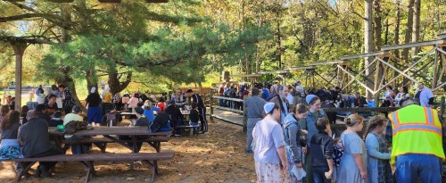 A large group of people gathered outdoors at picnic tables under trees, enjoying a community event in autumn.