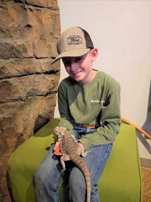 A young boy in a green shirt and cap sits on a green chair, holding a bearded dragon on his lap, smiling.