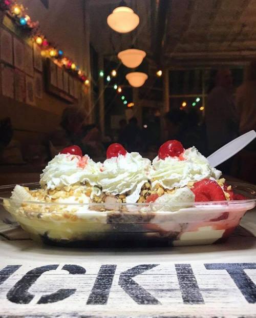 A colorful ice cream sundae topped with whipped cream, cherries, and nuts, served in a dish with a spoon.