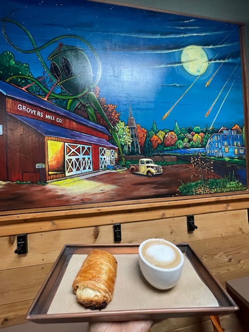 A hand holds a tray with a croissant and a small cup of coffee in front of a colorful mural depicting a night scene.