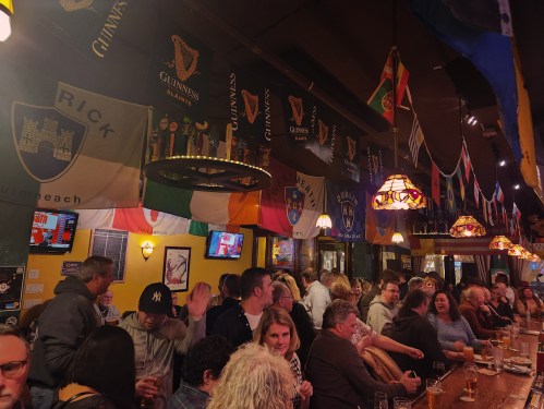 A lively bar scene with patrons enjoying drinks, surrounded by flags and Guinness banners. Warm, inviting atmosphere.