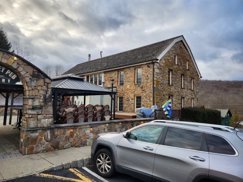 A stone building with a sloped roof, outdoor seating area, and a parked silver SUV in front. Cloudy sky in the background.