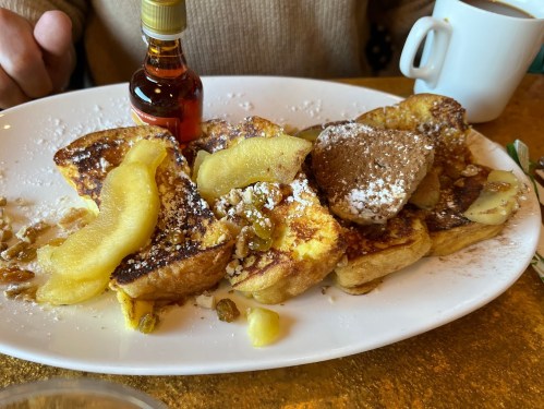 A plate of French toast topped with powdered sugar, apple slices, and a small bottle of syrup, with a coffee cup in the background.