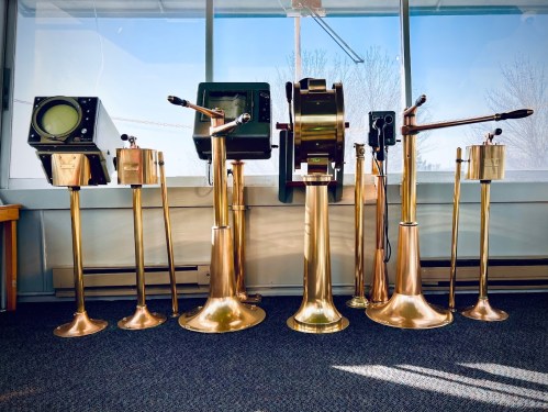 A row of vintage brass instruments and gauges displayed in a bright room with large windows.