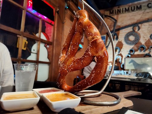 A large, golden-brown pretzel on a stand, served with three dipping sauces and a glass of water in the background.