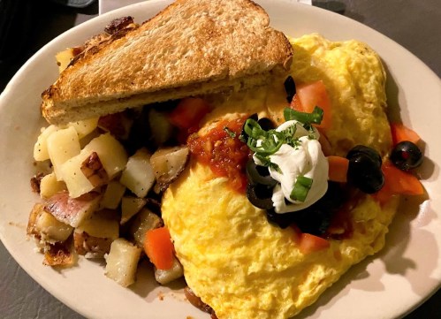 A plate of fluffy omelette topped with salsa, sour cream, olives, and tomatoes, served with toast and seasoned potatoes.