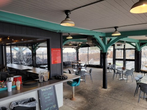 A cozy restaurant interior with teal accents, tables, and a bar area, featuring large windows and outdoor seating.