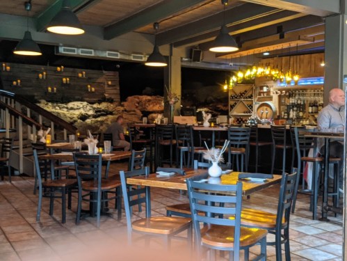 Cozy restaurant interior with wooden tables, black chairs, and a bar, featuring soft lighting and a stone wall backdrop.