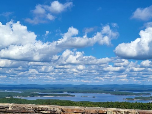 A scenic view of a lake surrounded by green hills and blue skies with fluffy clouds.