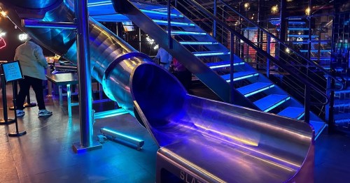 A metallic slide illuminated with blue lights, leading down from a staircase in a vibrant indoor space.