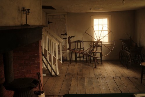 A rustic interior featuring a spinning wheel, wooden chairs, and a staircase, with soft light coming through a window.