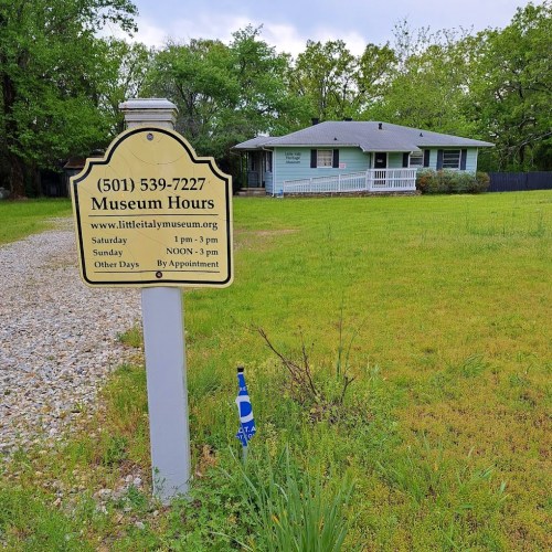 Sign displaying museum hours and contact information in front of a house surrounded by trees.