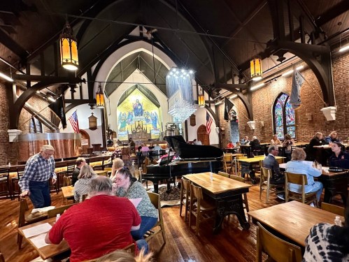 A lively restaurant interior with wooden tables, a grand piano, and a colorful mural, filled with diners and warm lighting.