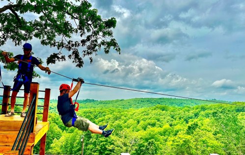 A person in a helmet zip lines over a lush green landscape, while an instructor assists from a platform.