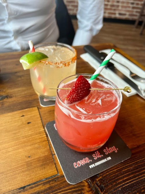 Two colorful cocktails on a wooden table, one garnished with a lime and the other with a strawberry.