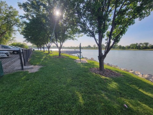 A serene riverside park with green grass, trees, and a bench, under a bright sun. A fence and vehicles are visible nearby.