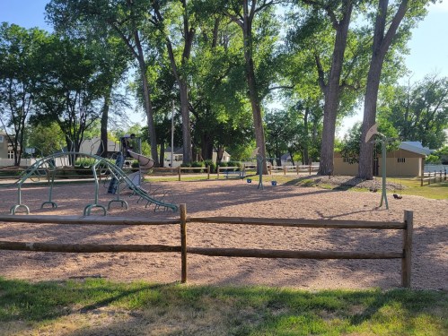 A sunny playground with slides and swings, surrounded by trees and a wooden fence.